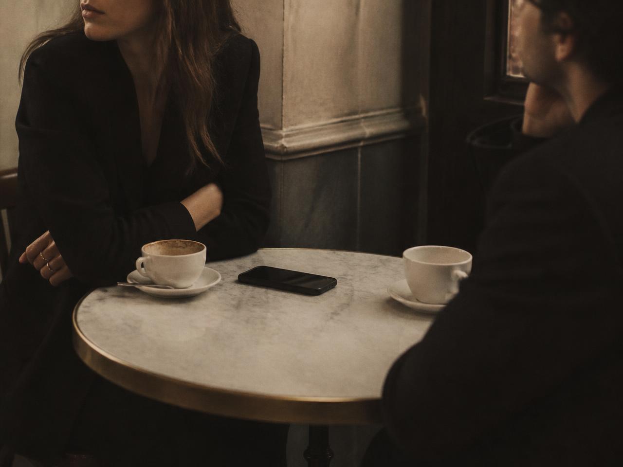 Two people on a first date at a cafe table, guarded body language, a phone face down between them